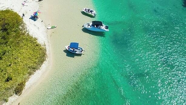 Eine Luftaufnahme eines Strandes mit klarem türkisblauem Wasser, einem sich ins Meer erstreckenden Steg und mehreren Booten, die in der Nähe des Ufers vor Anker liegen.