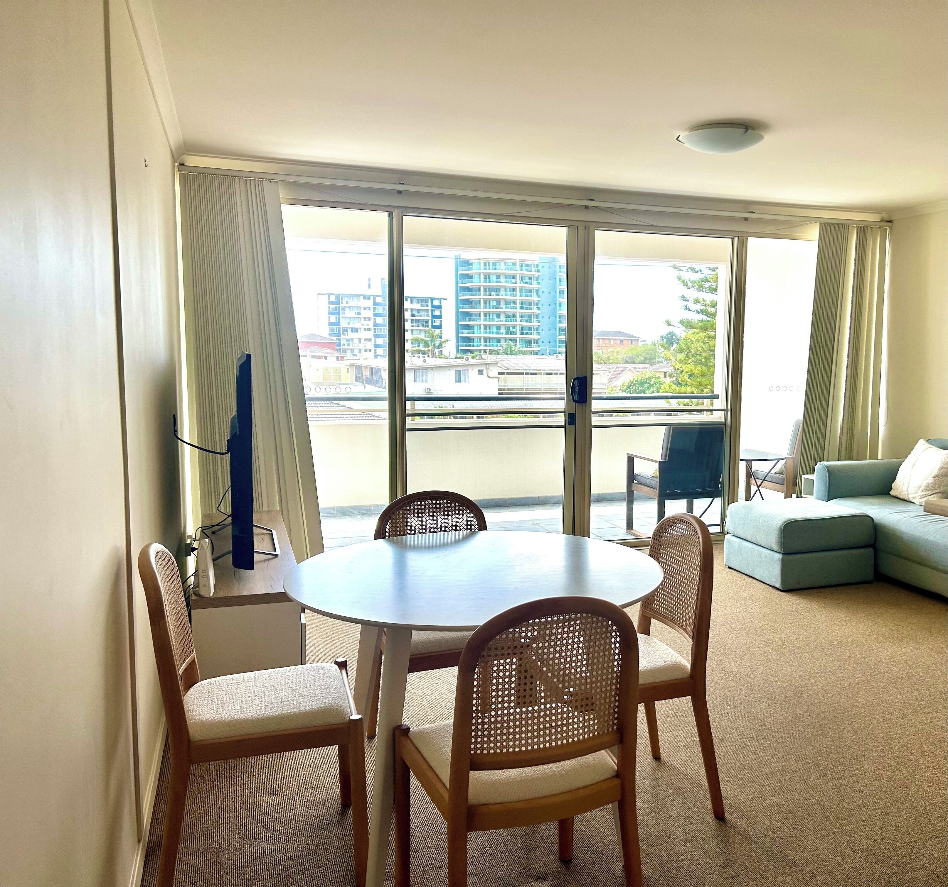 A room with a round table and four chairs, a sofa, and a large window with a view of buildings.