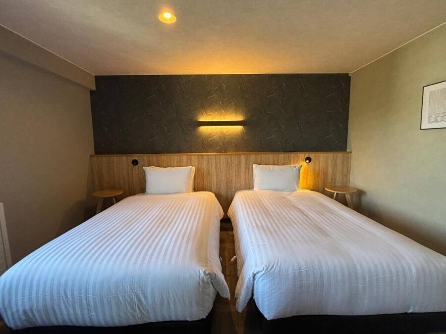 A hotel room with two single beds, a dark textured wall, and a wall-mounted light fixture.