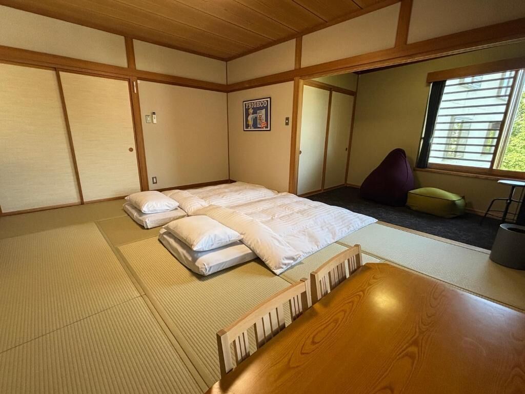 A traditional Japanese room with tatami flooring, a futon bed, a wooden table, and a window with blinds.