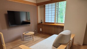 Ein Zimmer mit einem Bett, einem an der Wand befestigten Fernseher, einem kleinen Tisch und einem Fenster mit Blick auf Bäume.