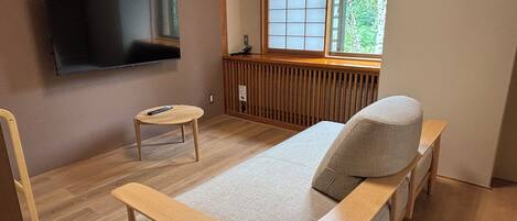 Una habitación con una cama, televisión montada en la pared, una mesita y ventana con vistas a árboles.