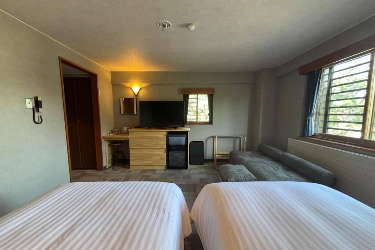 Habitación de hotel con dos camas, un escritorio con televisor, una pequeña kitchenette y una ventana con persianas.
