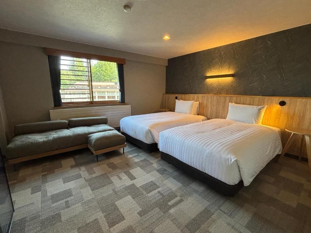 Une chambre d’hôtel avec deux lits, un canapé, une fenêtre avec des stores et un luminaire fixé au mur.