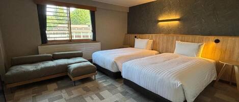 Une chambre d’hôtel avec deux lits, un canapé, une fenêtre avec des stores et un luminaire fixé au mur.