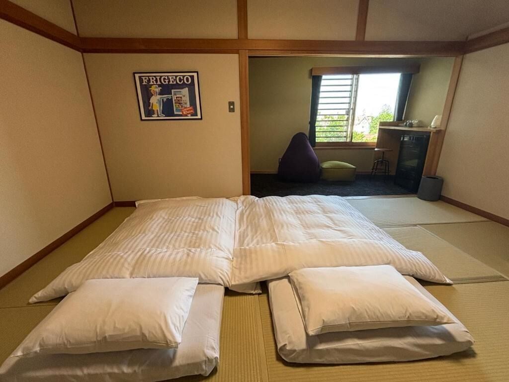 A room with tatami flooring, a bed with white bedding, a small table, and a chair.