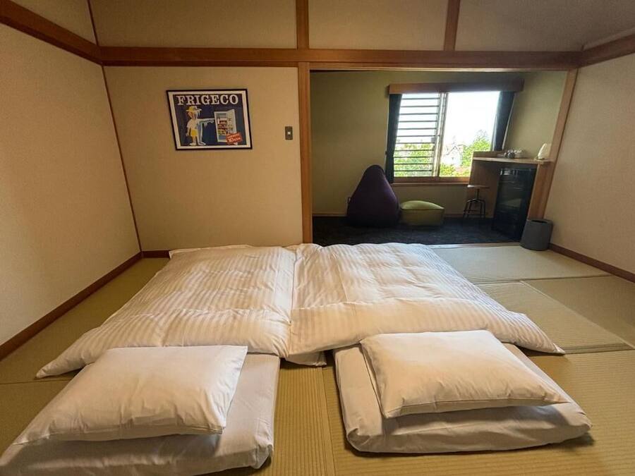 Una habitación con suelo de tatami, una cama con ropa de cama blanca, una mesita y una silla.