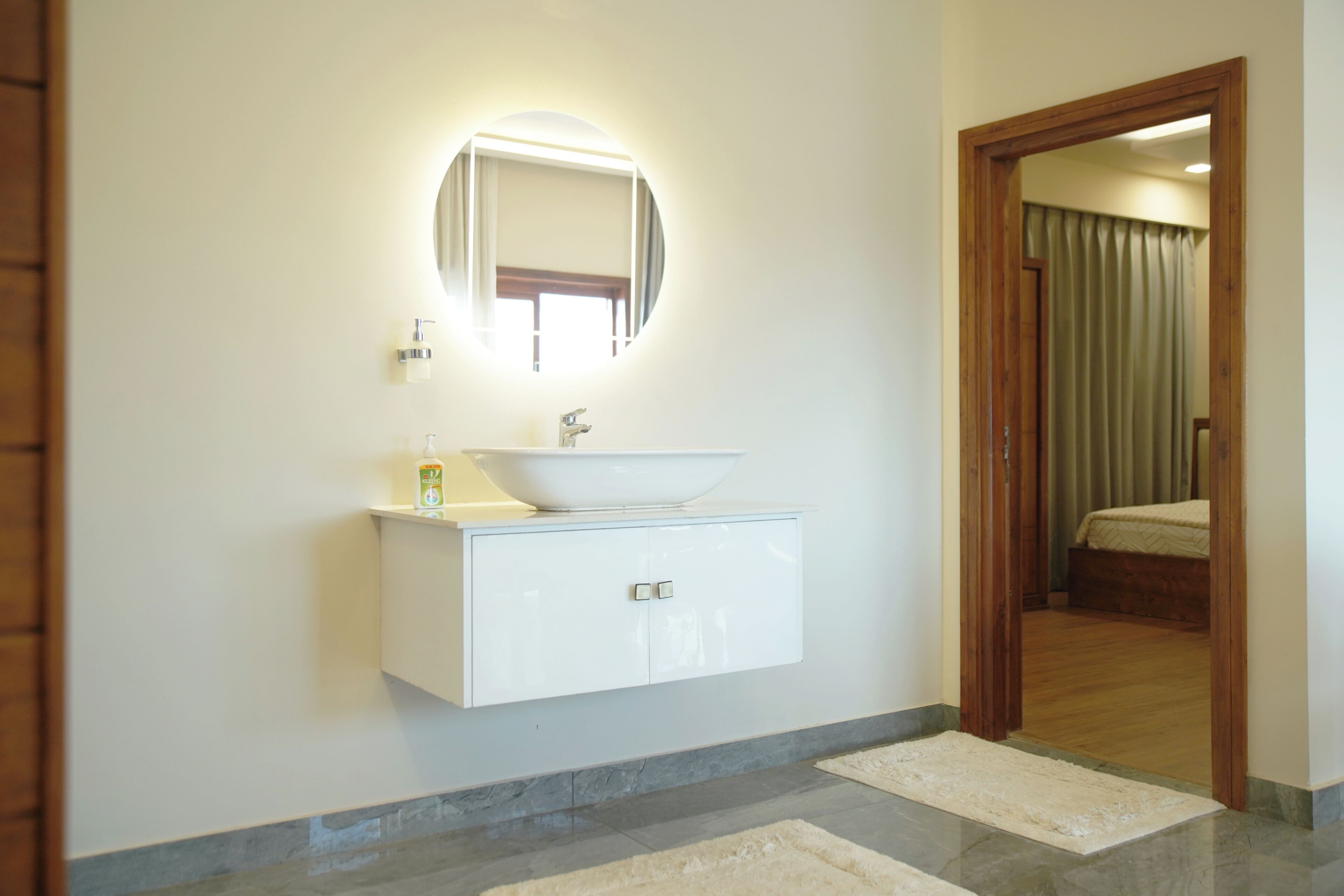 A modern bathroom with a white vanity, a round mirror with lighting, and a view into a bedroom through an open door.