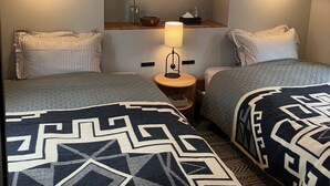 Quarto de hotel com duas camas, uma mesinha de cabeceira com abajur, um pequeno armário com artigos de higiene pessoal e um detalhe no teto em design ripado.
