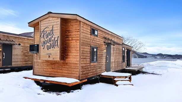 Uma cabana com fachada de madeira, uma placa com a inscrição 'Encontre sua essência selvagem' e uma pequena área de deck.