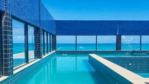 A swimming pool with a view of the ocean, surrounded by glass walls and wooden flooring. - Ocean View on the 20th Floor | Rooftop with Jacuzzi | 1 Block from the Beach | (Maceió)