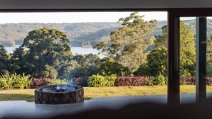 A room with a stone fire pit and a view of a lake surrounded by trees.