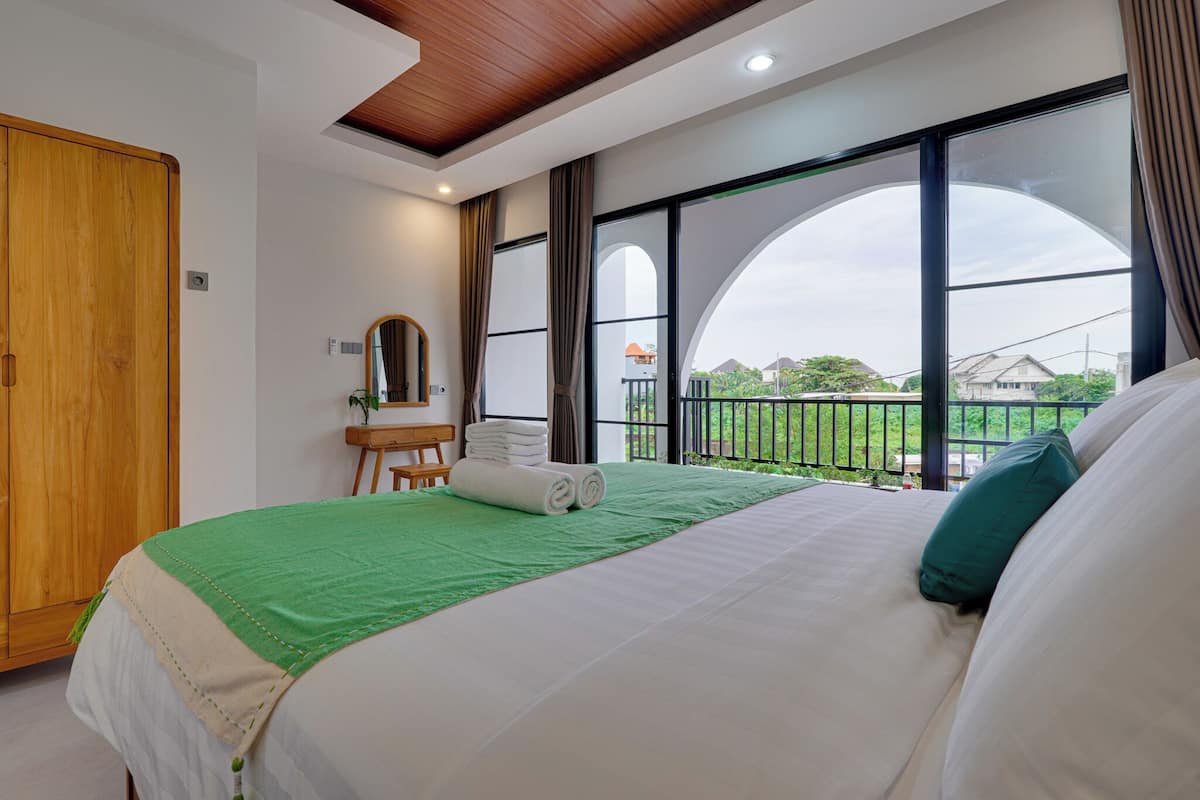 Une chambre d’hôtel avec un grand lit, une armoire en bois, un bureau avec un miroir et une vue sur l’extérieur grâce à de grandes fenêtres.