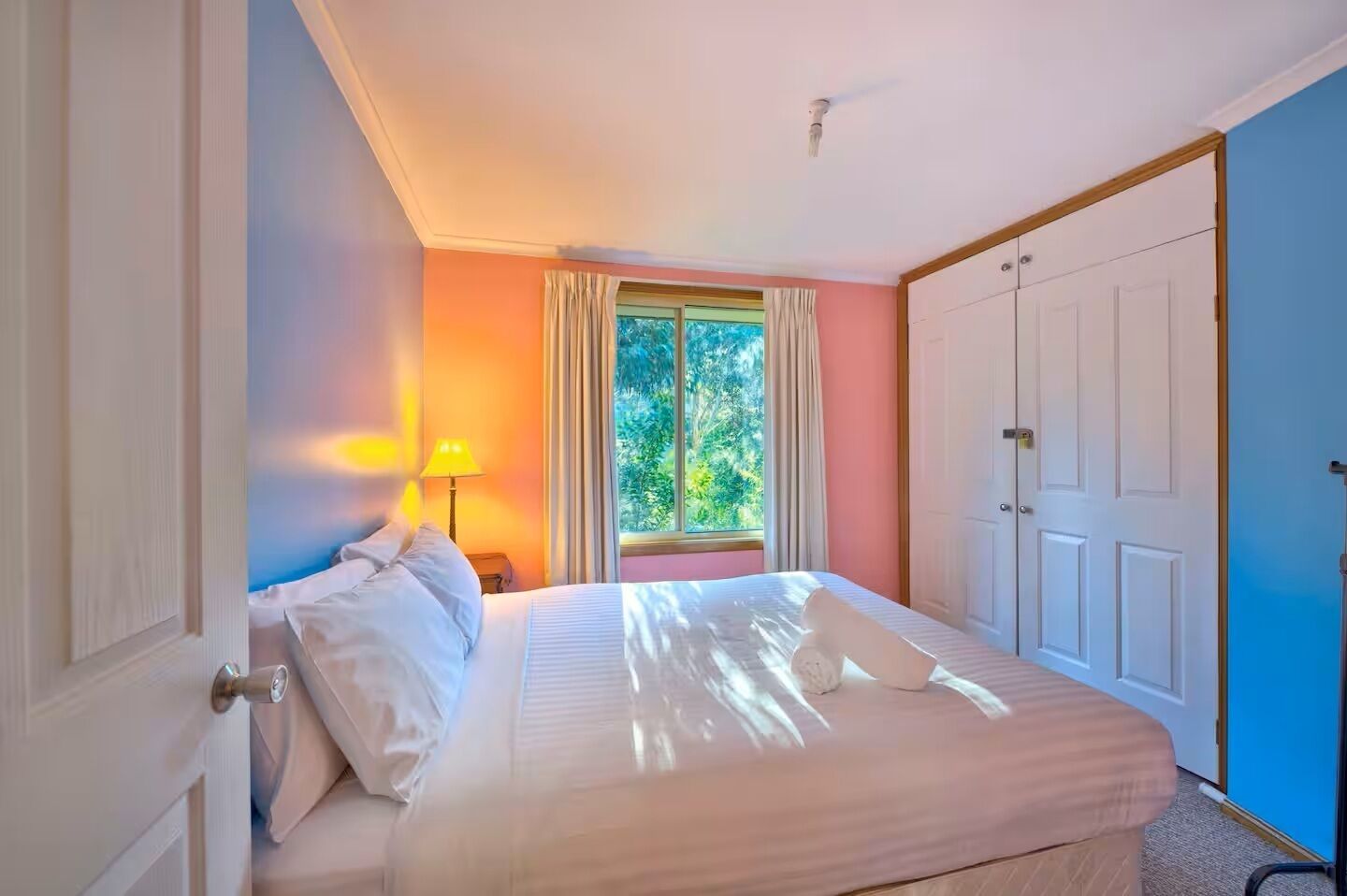 A bedroom with a bed, a window with curtains, a lamp, and a wardrobe.