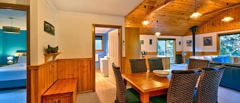 A cozy room with a wooden dining table, wicker chairs, and a bench. There is a window with a view of trees and a door leading to another room.