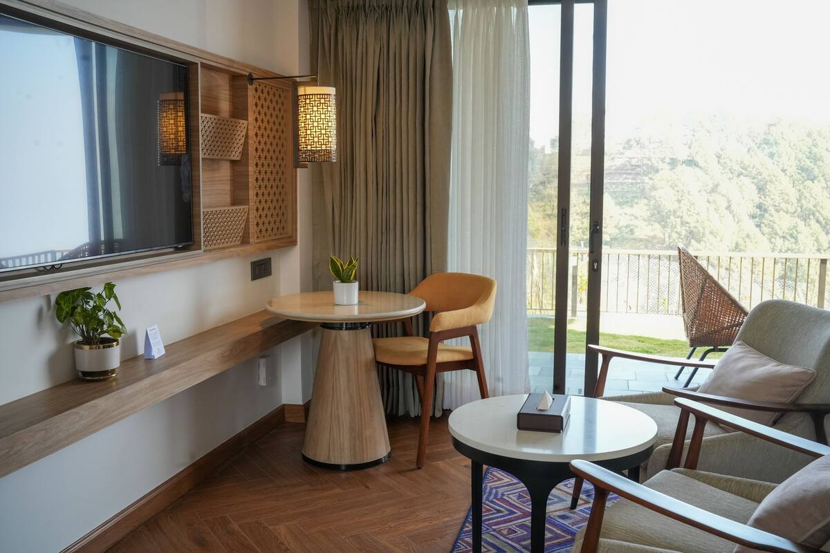 A balcony with a wooden table, chairs, a potted plant, and a view of a mountainous landscape.