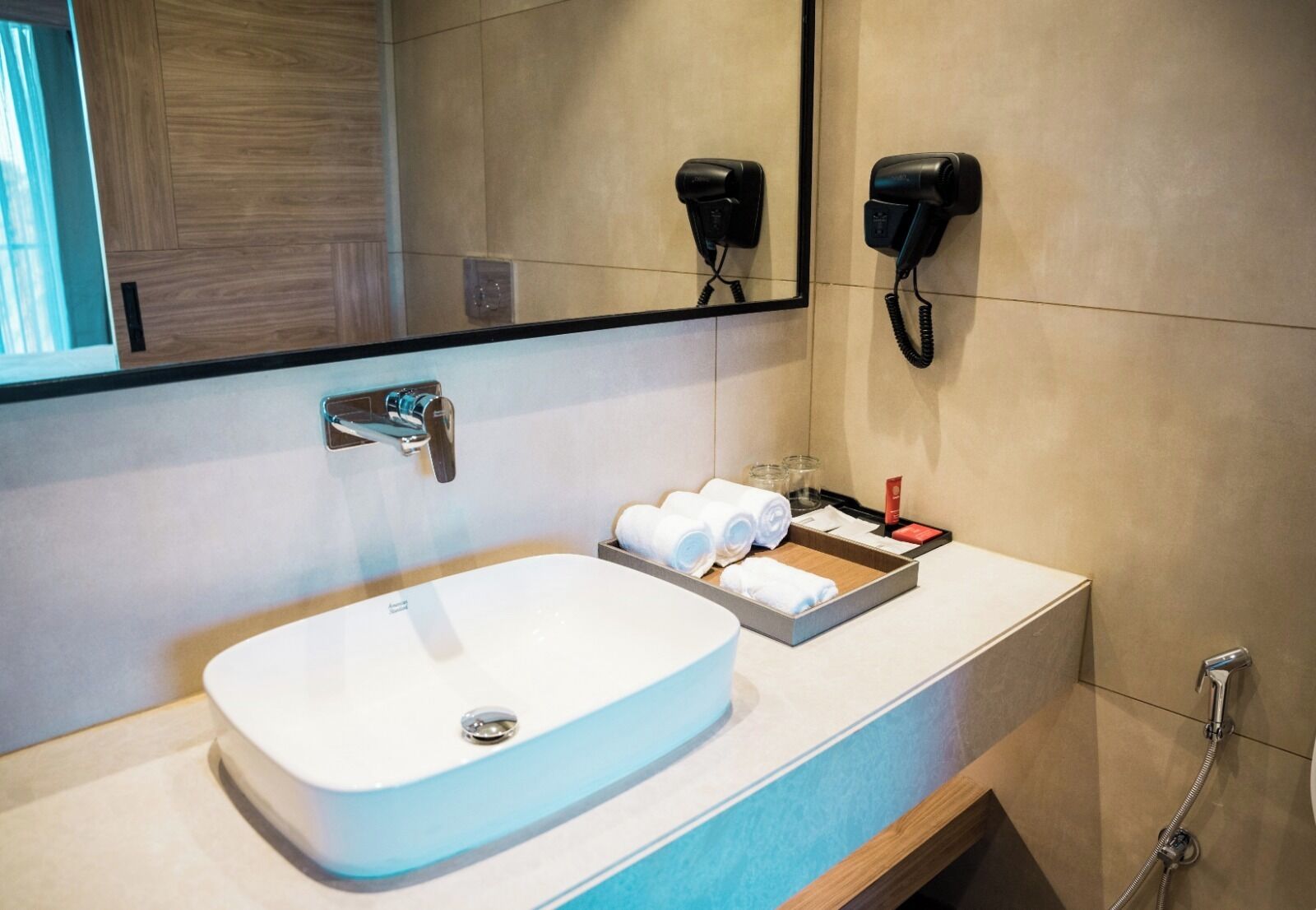 Casa de banho moderna com lavatório branco, espelho, dois secadores de cabelo e um prato com toalhas e produtos de higiene pessoal.
