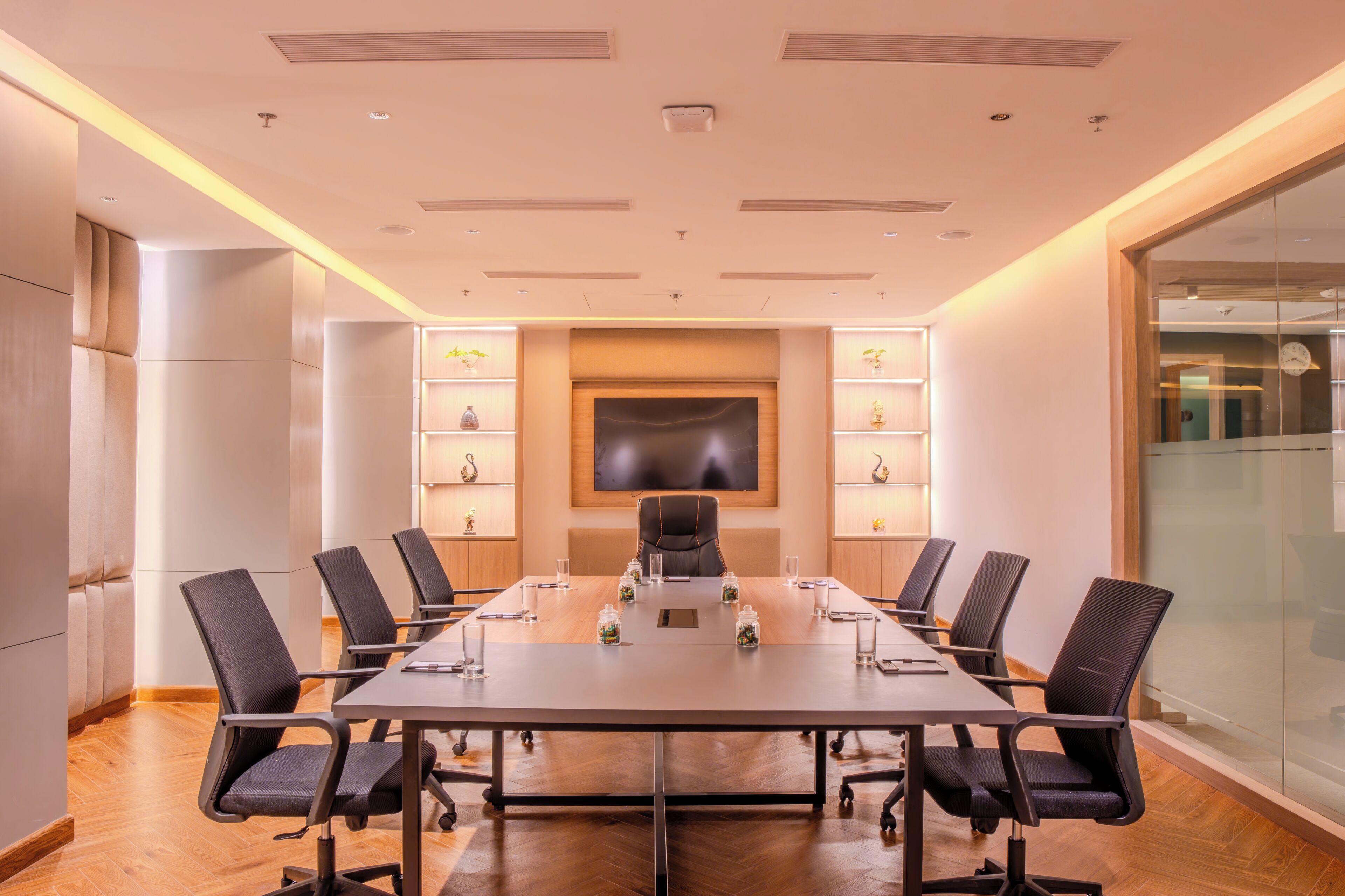 A conference room with a long table, black chairs, a flat-screen TV, and shelves with decorative items.
