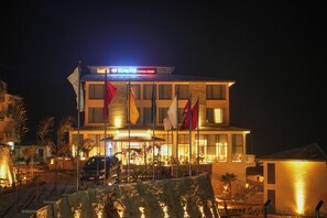 Un hôtel dont l’entrée est bien éclairée, qui arbore plusieurs drapeaux et devant lequel une voiture est visiblement garée.