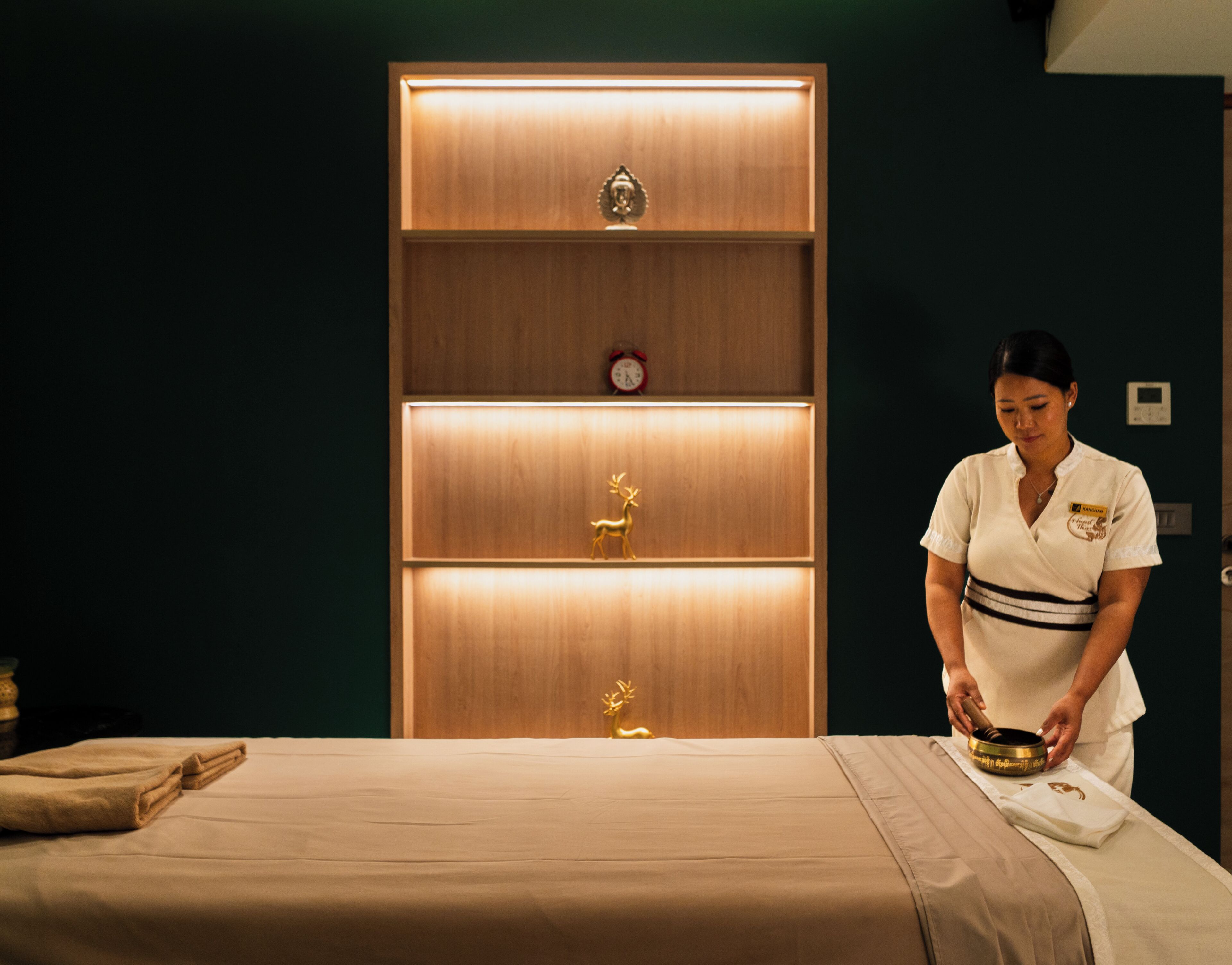 A room with a bed, a wooden cabinet with shelves, and a person preparing a massage table.