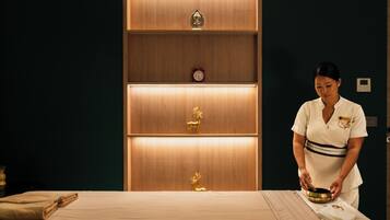 A room with a bed, a wooden cabinet with shelves, and a person preparing a massage table.