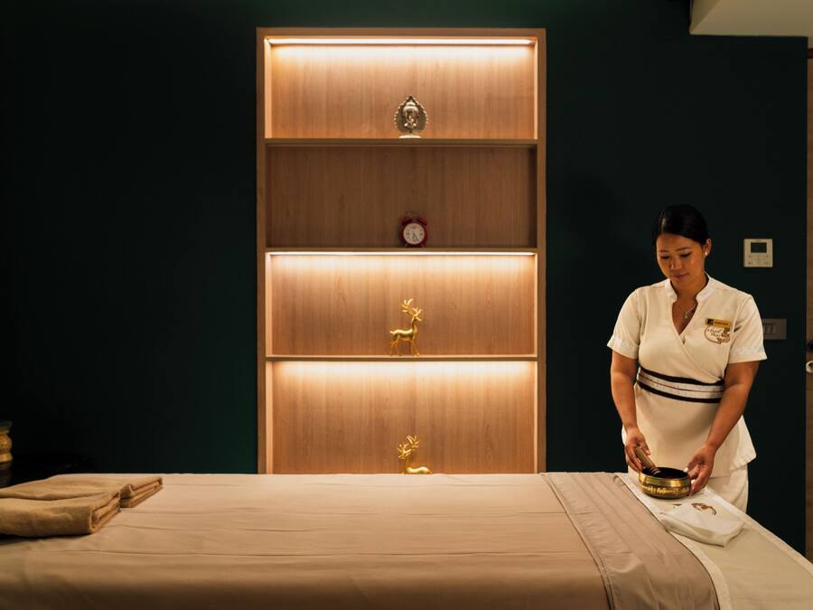 A room with a bed, a wooden cabinet with shelves, and a person preparing a massage table.