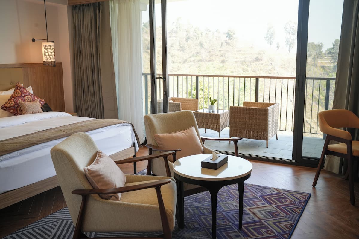 A hotel room with a bed, two armchairs, a small table, and a balcony with wicker furniture.