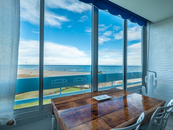 A room with a wooden table and chairs, large windows with a view of the beach and ocean, and a blue railing along the window. - The Big Blue! Between sea and mountains (Saint-Cyprien)