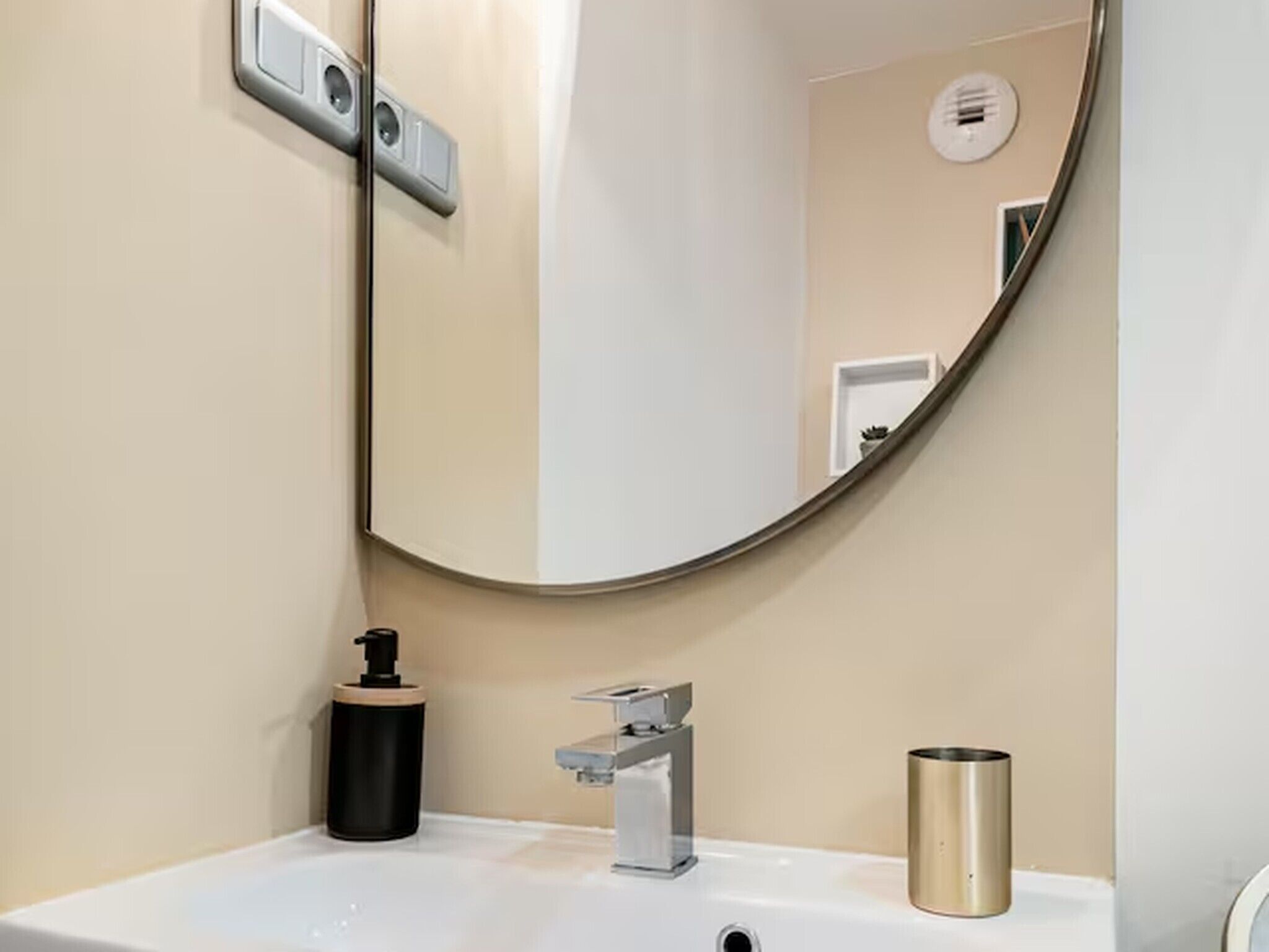 A bathroom with a round mirror, a sink, a faucet, a soap dispenser, and a trash can.