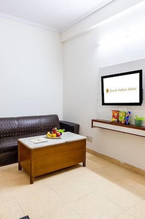 Hotelkamer met een houten salontafel, een donkerbruine bank, een flatscreen televisie aan de muur en een plank met snacks en een potplant.