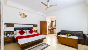 A hotel room with a bed, a wooden nightstand, a desk, a brown leather sofa, a ceiling fan, and a dining area with a chair and table. - Trevon Inn Karol Bagh, New Delhi (New Delhi)
