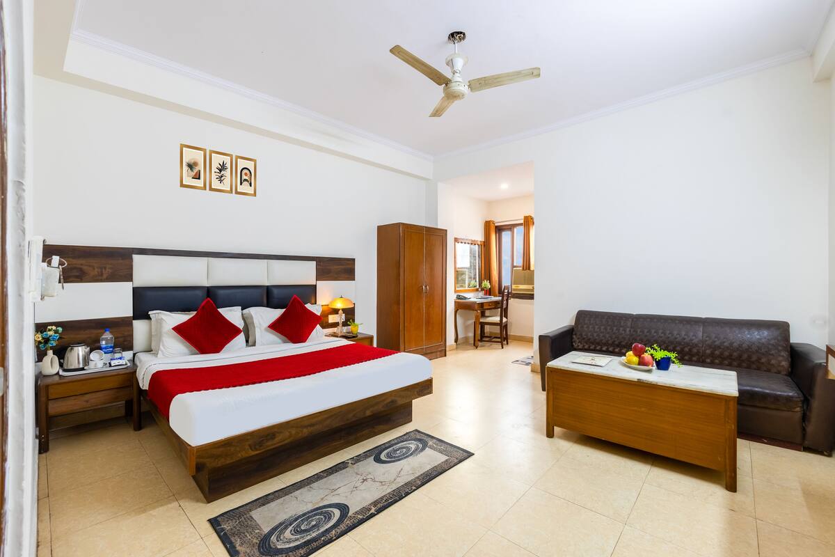 Quarto de hotel com cama, mesa de cabeceira de madeira, escrivaninha, sofá de couro marrom, ventoinha no teto e área de jantar com cadeira e mesa.