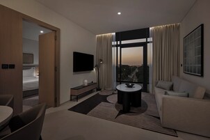 Habitación de hotel moderna con un ventanal grande, televisor de pantalla plana, sofá, mesa de comedor y una mesita auxiliar.