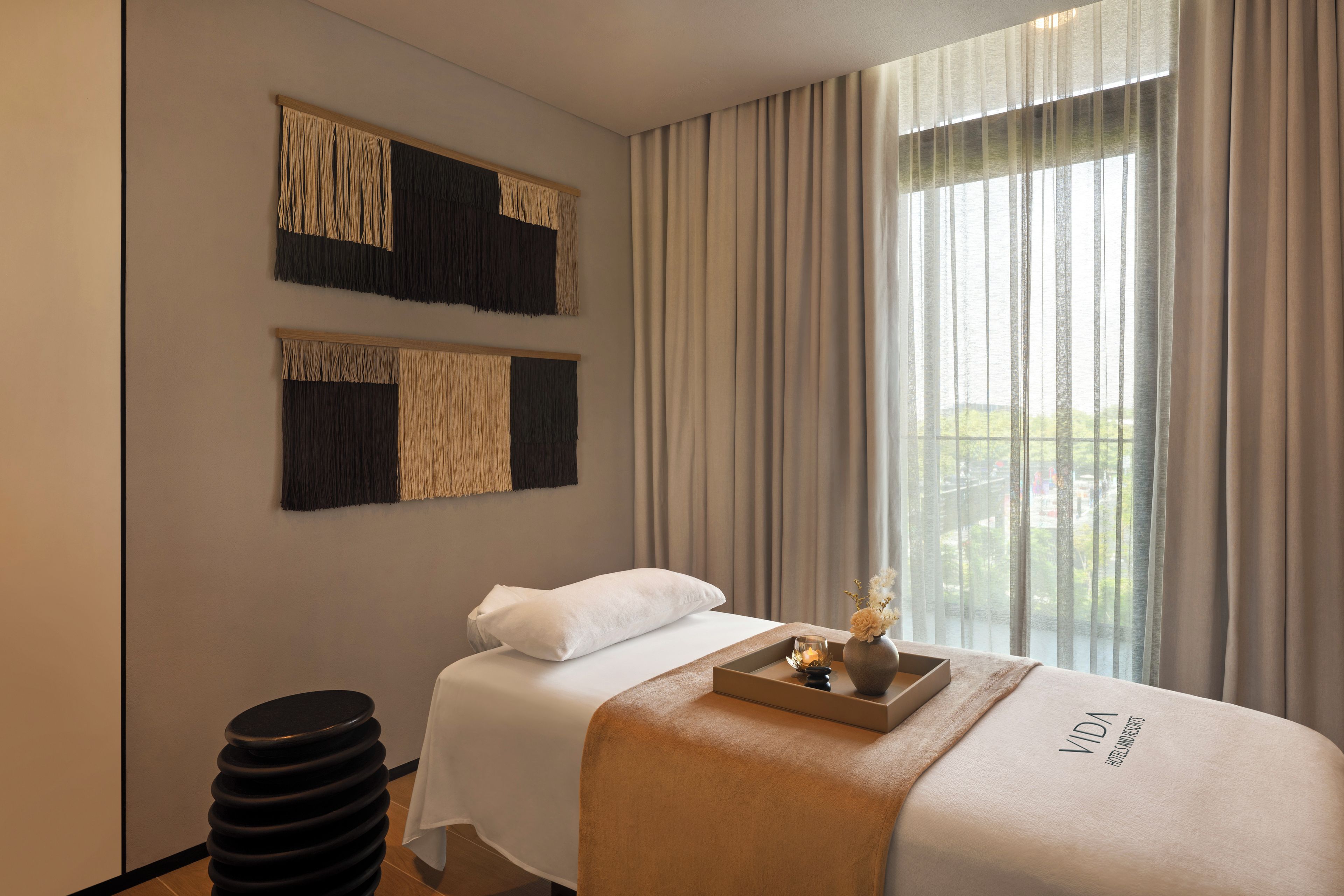 A hotel room with a bed, a bedside table with a vase, and a window with curtains.