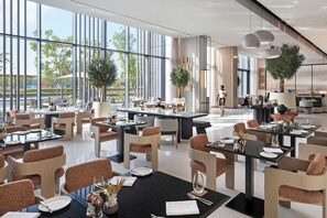 A modern restaurant with large windows, multiple dining areas, and indoor plants. - Vida Aljada (Sharjah)