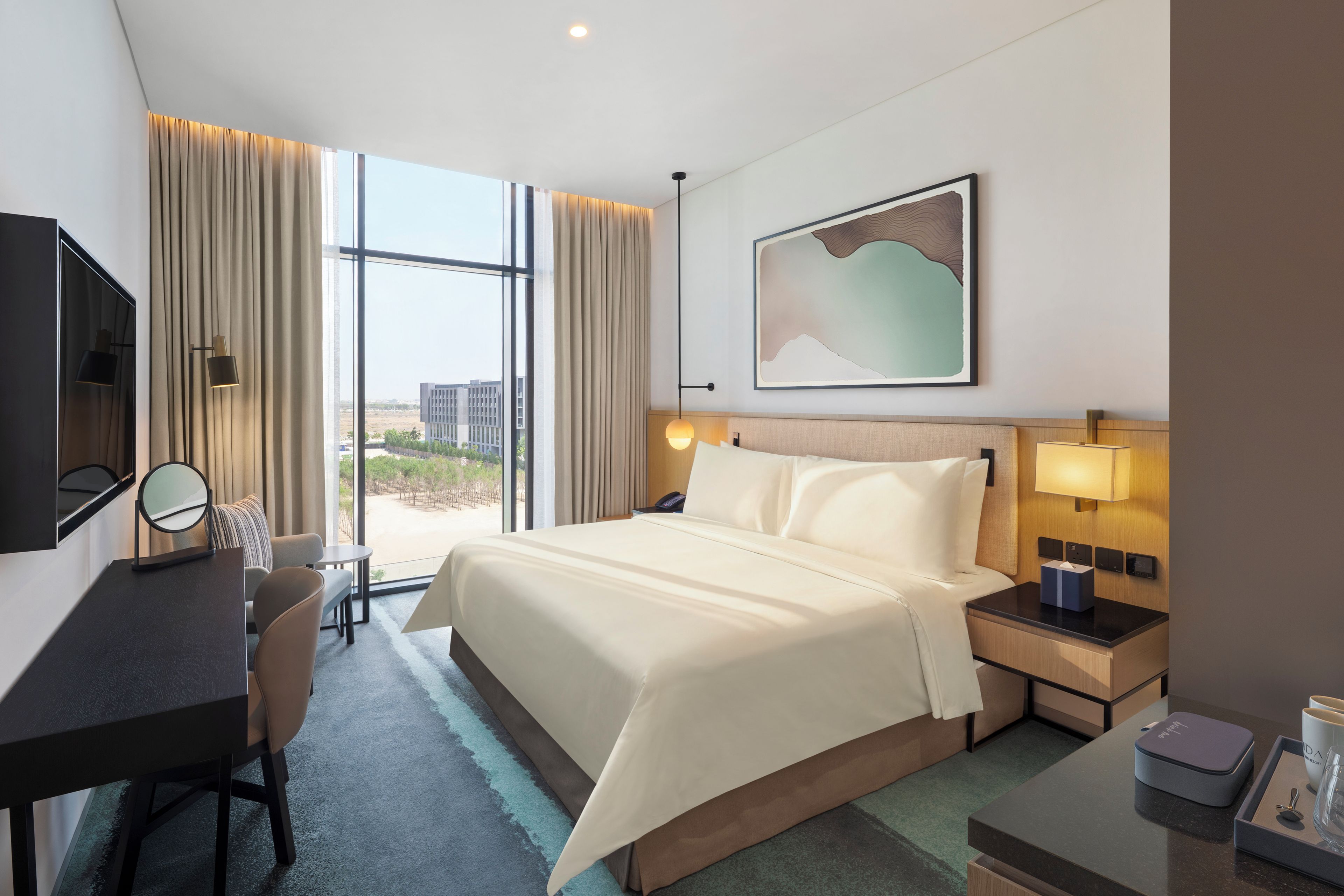 Quarto de hotel com uma cama grande, uma escrivaninha com cadeira, mesa de cabeceira com abajur e vista para o exterior.
