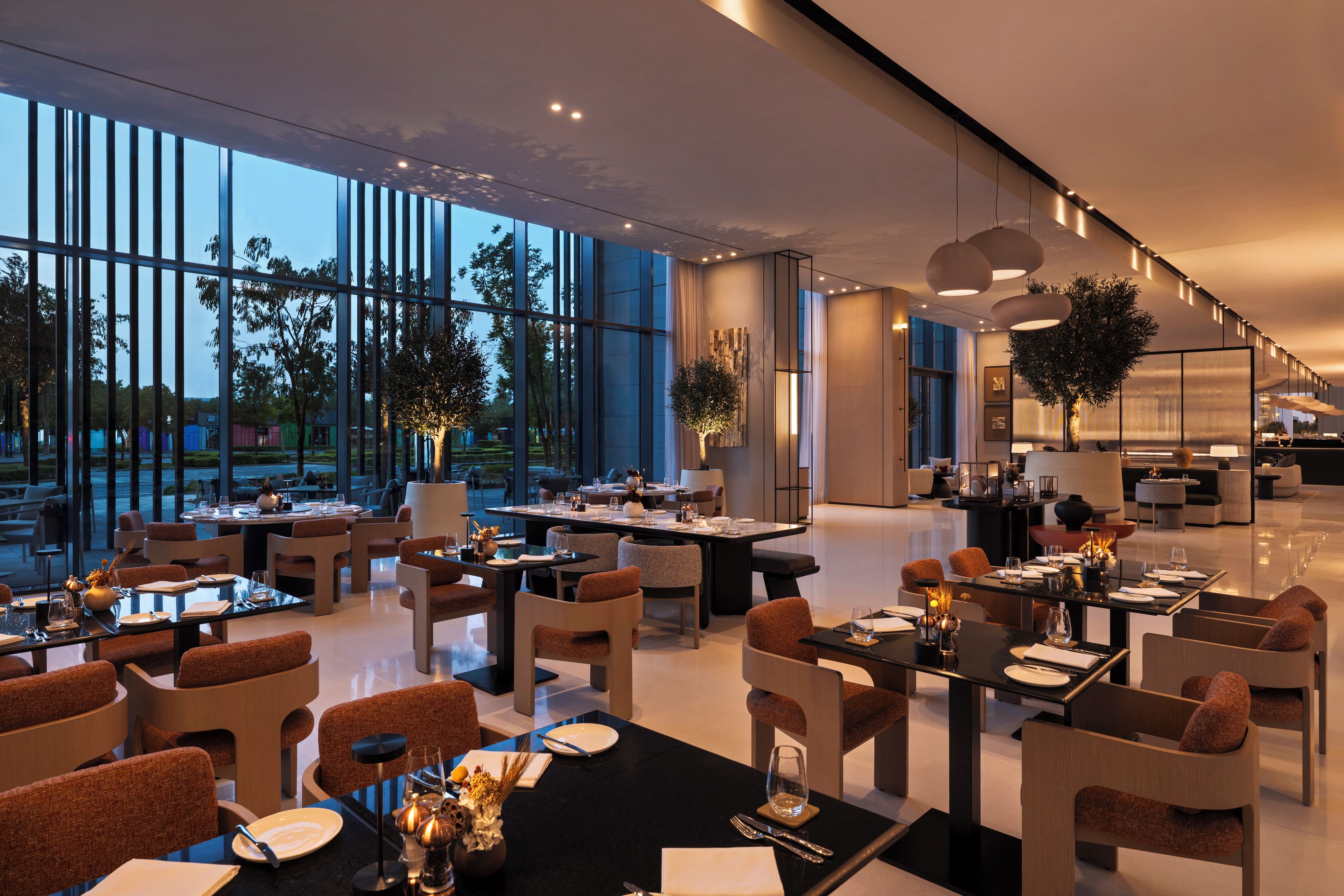 Un espace de restauration moderne dans un hôtel, avec de nombreuses tables et chaises, de grandes fenêtres et des plantes d’intérieur.