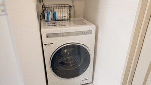 Máquina de lavar roupa de carregamento frontal, compacta e branca, num canto do quarto, com parede branca texturizada e moldura de porta visíveis.