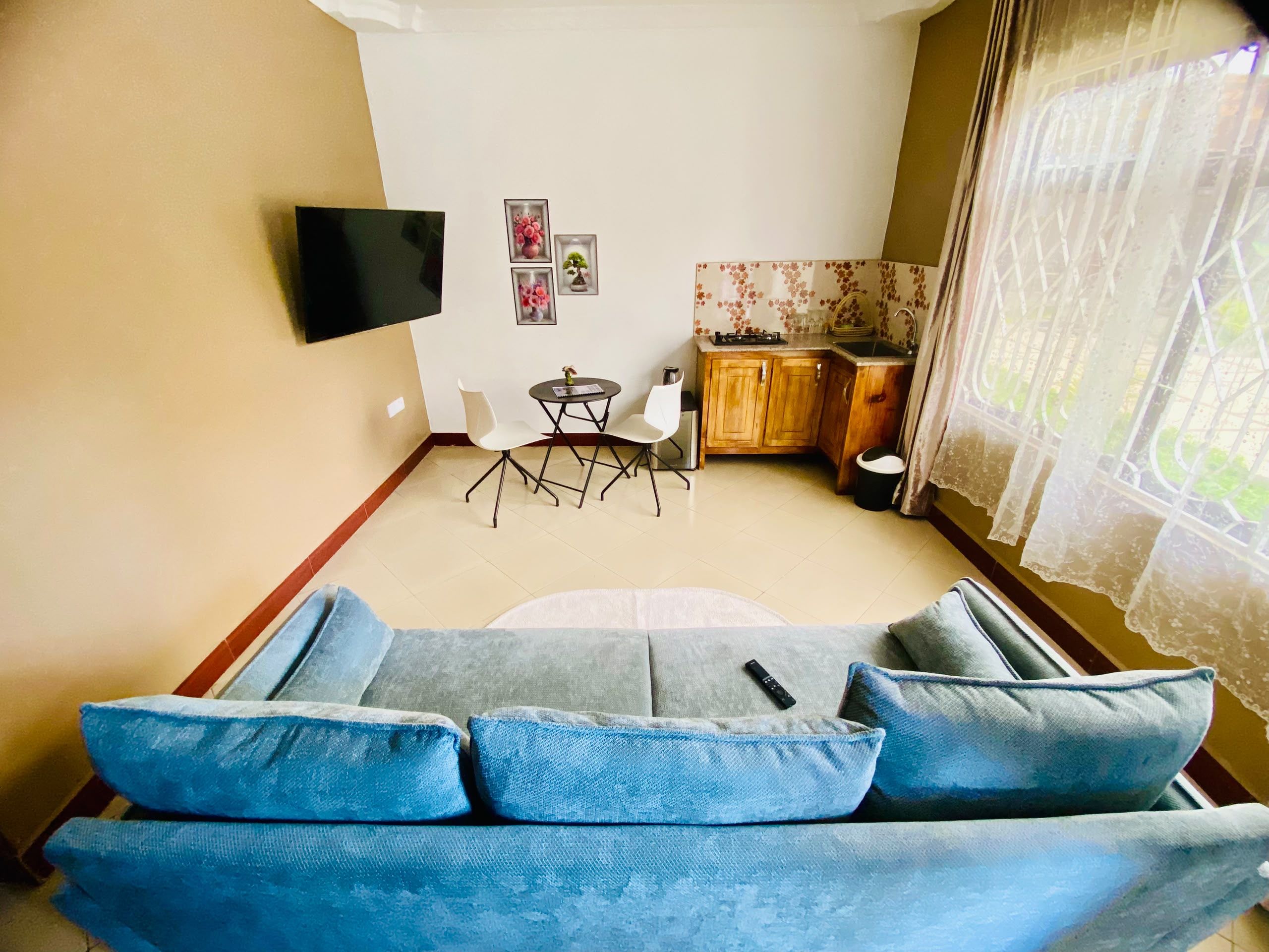 Ein Wohnzimmer mit einer Couch, einem an der Wand befestigten Fernseher, einem Essbereich mit Stühlen, einer kleinen Küche mit Schränken und einem Fenster mit Vorhängen.
