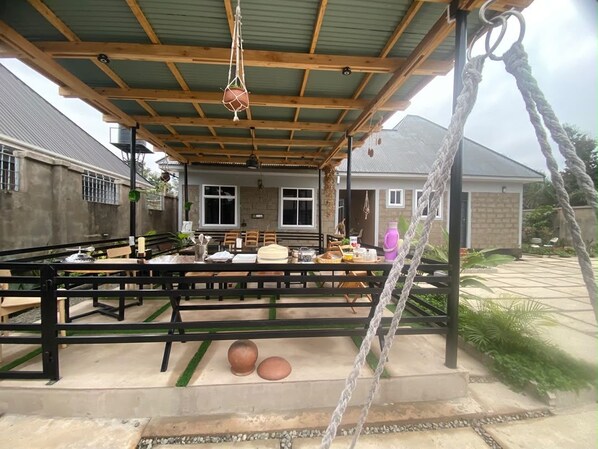 Un área de comedor al aire libre con techo cubierto, mesas, sillas y un columpio de cuerda.