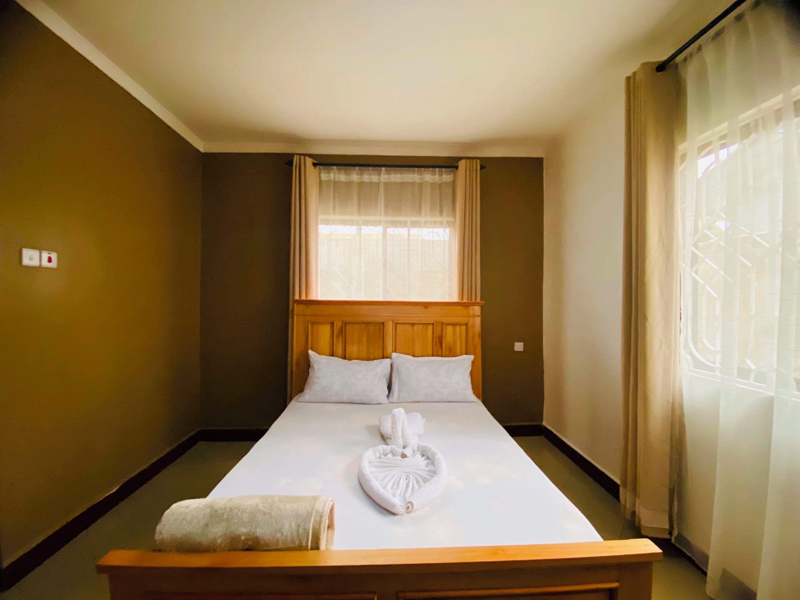 Ein Hotelzimmer mit einem Bett, einem Holzkopfstück, einem Fenster mit Vorhängen und einem Handtuch auf dem Bett.