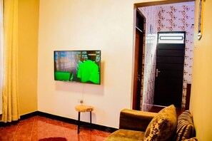 A living room with a flat-screen TV mounted on the wall, a small stool, and a sofa with patterned cushions. -  Hideaway Near Kilimanjaro Airport Private Pool & Chemka Hot Springs Access (Boma Ng'ombe)