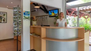 A reception desk with a mounted television, a postcard rack, and a large window with a view of greenery outside. - Cottage Dourbie 4p 2bdrms Aircon (Millau)