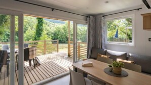 A modern room with a wooden deck, a dining area with a table and chairs, and a large window offering a view of greenery. - Cottage Dourbie 4p 2bdrms Aircon (Millau)