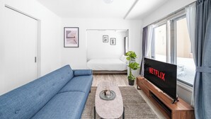 A modern living room with a blue sofa, a coffee table, a TV displaying 'NETFLIX', and a large window with curtains. - VOLPE 102 (Sapporo)