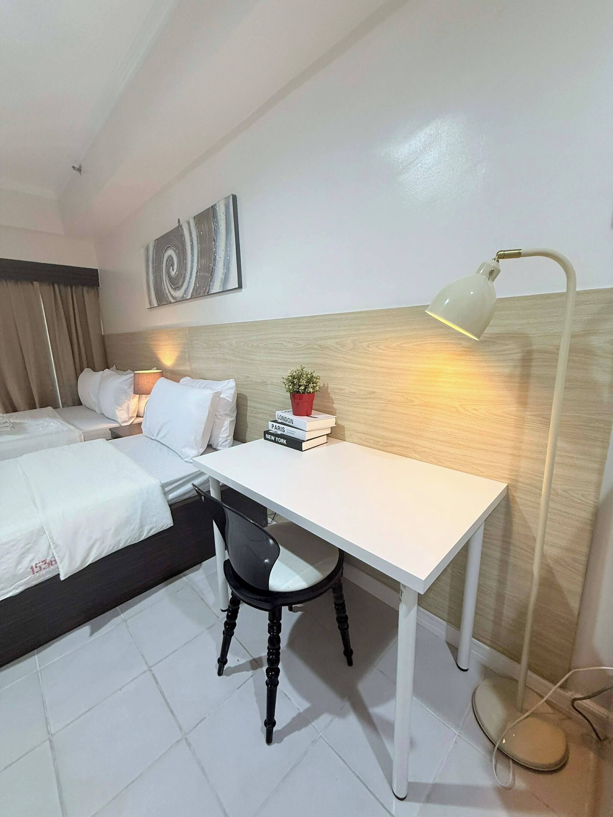 Habitación de hotel con cama, escritorio, silla, lámpara, una obra de arte en la pared y una pequeña planta en maceta.