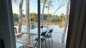 Een glazen terras met een tafel en stoelen, uitkijkend op een rustig wateroppervlak en bomen.