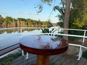 Une terrasse en bois avec une table ronde et une balustrade blanche, offrant une vue sur un lac paisible et une végétation luxuriante.