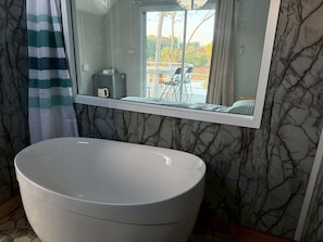 Une salle de bain dotée d’une grande baignoire sur pieds, d’un mur au motif de marbre, d’un rideau de douche à rayures et d’un miroir qui reflète un balcon avec des meubles d’extérieur.