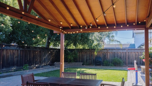 A covered patio with a wooden table and chairs, a basketball hoop, and a wooden fence in the background.
